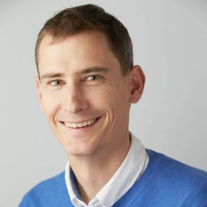 Dr Nicholas McLernon smiles at the camera in front of a white background. He is wearing a white shit with a blue jumper on top